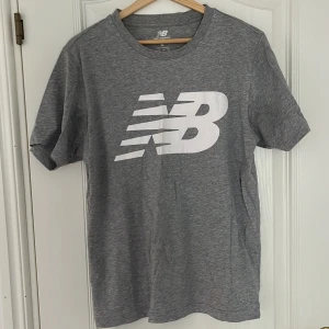 Grå t-shirt - New Balance - Säljer en grå t-shirt från New Balance med stor vit logga framtill. Klassisk passform och rund halsringning. Perfekt för träning eller vardag. Mjuk och bekväm bomullskänsla.