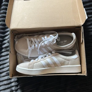 Vita Adidas Campus sneakers - Säljer ett par vita Adidas Campus sneakers med klassiska tre ränder på sidan. Skorna har snörning, rund tå och är tillverkade i mocka och textil. Perfekta för en stilren och sportig look och passar perfekt nu till sommaren. Den högra skol har en lite fläck men den syns knappt☺️storleken är ungefär 39. Det är köpta på Sellpy för 700kr!!