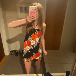 Svart kortklänning med blommigt mönster - Säljer en svart kortklänning med färgglada blommor i rött, orange och vitt samt gröna blad. Klänningen är ärmlös med smala axelband och har en lätt volang nertill. Perfekt för varma dagar och somriga tillfällen. Klänningen är gammal so vet inte vad det är för storlek eftersom det inte står någon lapp 