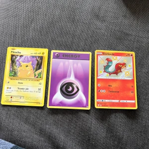 Pokémonkort: Pikachu, Psychic Energy, Sizzlipede - Tre Pokémonkort säljes: Pikachu (gul med lila bakgrund), Psychic Energy (lila med öga-symbol) och Sizzlipede (röd med blå och orange detaljer). Samlarkort med klassisk Pokémon-design och färgglada illustrationer.