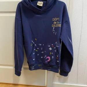 Gallery Dept Hoodie - Säljer en blå hoodie från Gallery Dept. med trycket 'DÉPT. de la GALERIE' på bröstet och färgglada penseldrag över hela tröjan. Hoodien har känguruficka och en avslappnad passform. Perfekt för dig som gillar streetwear med unik design. Storlek S baggy fit. ❗️OBS PÅSEN MEDFÖLJER INTE❗️
