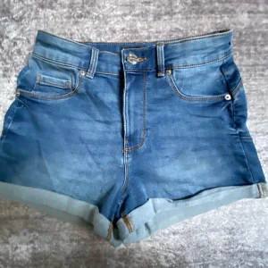 Blå jeansshorts från FB Sister/New Yorker  - Snygga blå jeansshorts från FB Sister med klassisk femficksdesign och uppvikta benslut. Shortsen har normal passform och stängs med knapp och dragkedja. Perfekta för varma dagar och enkla att matcha med olika toppar.