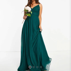 Mörkgrön långklänning med rosett - Elegant mörkgrön långklänning med smala axelband och v-ringad omlott-topp. Klänningen har en stor rosett i ryggen och ett fint fall i kjolen. Perfekt för bal eller festliga tillfällen. Man kan även knyta banden lite hur man vill:) Har använt 1 gång. 