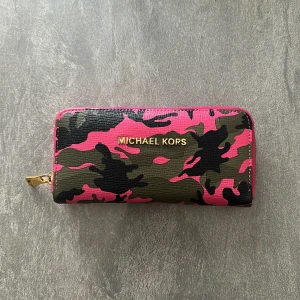 Michael Kors plånbok med camouflage - Cool plånbok från Michael Kors i rosa och grönt camouflage-mönster med guldfärgad dragkedja. Plånboken har flera kortfack och ett innerfack med dragkedja. Perfekt för dig som vill sticka ut med din accessoar.