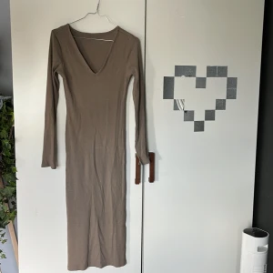 Brun ribbad långklänning från 157 - Säljer en brun ribbad långklänning från 157 med v-ringning och långa ärmar. Klänningen har en tight passform och är tillverkad i ett mjukt, stretchigt material som sitter snyggt på kroppen. Perfekt för dig som gillar stilrena och enkla plagg.