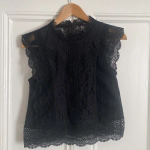 Fest topp - Säljer en svart blus i spets med fina volangdetaljer längs ärmar och nederkant. Blusen är ärmlös och har rund halsringning samt dragkedja i ryggen. Perfekt för dig som gillar romantisk och elegant stil.