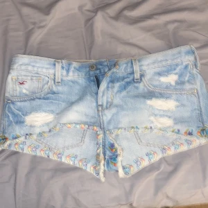 Ljusblå jeansshorts med broderi från Hollister - Säljer ett par ljusblå jeansshorts från Hollister med slitna detaljer och färgglatt broderi längs bensluten. Shortsen har fransig kant, klassiska fickor och låg midja. Perfekta för sommaren! Knappt andvänd mellan storlek s till m ❤️