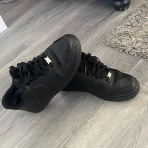 Svarta Nike Air Force 1 sneakers - Säljer ett par helsvarta Nike Air Force 1 sneakers med klassisk design. Skorna har snörning, rund tå och platt sula. Perfekta för dig som gillar stilrena och tidlösa sneakers.