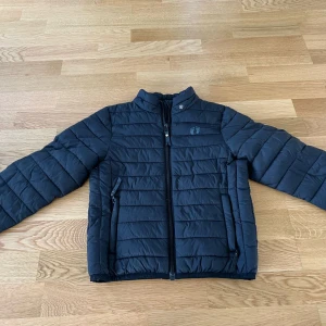 Svart dunjacka från Fjällräven - Säljer en svart dunjacka från Fjällräven med ståkrage och dragkedja framtill. Jackan har två sidofickor med dragkedja och diskret logga på bröstet. Perfekt för kyliga dagar och enkel att matcha med olika outfits.