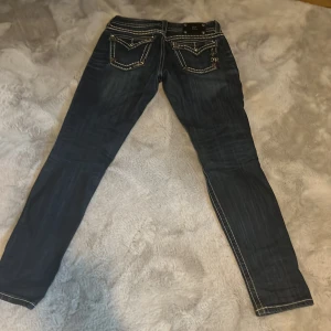 Mörkblå jeansbyxor från Miss Me - Säljer ett par mörkblå jeans från Miss Me med snygga kontrastsömmar och broderade detaljer på bakfickorna. Byxorna har normal passform och dragkedja framtill. Perfekta för dig som gillar jeans med unika detaljer.