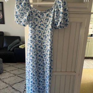 Blommig långklänning från Gina Tricot - Superfin långklänning från Gina Tricot med blått blommönster på vit botten. Klänningen har puffiga korta ärmar och knytning framtill vid halsen. Perfekt för sommarens alla tillfällen!