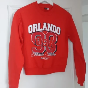 Röd sweatshirt med Orlando-tryck - Röd sweatshirt med stort tryck framtill: 'Orlando 98 USA Florida America Sport' i vitt, blått och rött. Rund halsringning och ribbade muddar vid ärmslut och nederkant. Perfekt för en sportig look.