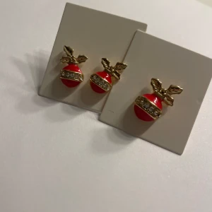 Juliga örhängen med röd och guld dekor - Säljer ett par örhängen formade som julkulor i rött och guld med små glittrande stenar och dekorativa blad upptill. Perfekta för att addera lite extra festlig känsla till din look!