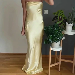 Ljusgul balklänning satin - Säljer en elegant ljusgul långklänning i satin med axelbandslös design och öppen rygg. Klänningen har en slät, figurnära passform och faller snyggt längs kroppen. Perfekt för bal eller student.