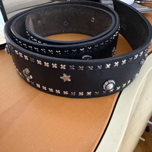 Svart bälte med nitar och stjärndetaljer - Säljer ett svart bälte med silvriga nitar och stjärnformade detaljer. Bältet har även runda dekorationer och passar perfekt till jeans. Snygg accessoar för att ge outfiten lite edge.