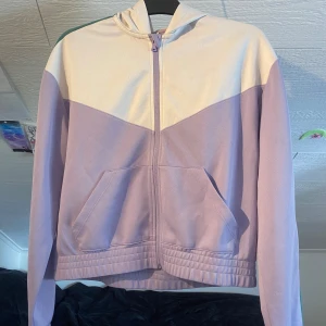 Ljuslila och vit hoodie från H&M Move - Säljer en hoodie från H&M Move i ljuslila och vitt med dragkedja framtill. Jackan har huva, ribbade muddar och fickor på magen. Perfekt för en sportig och avslappnad stil.