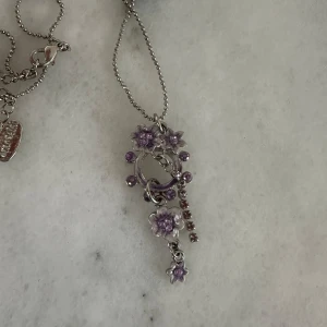 Silverfärgat halsband med lila blommor - Säljer ett silverfärgat halsband med hänge dekorerat med lila blommor och små glittriga stenar. Kedjan är tunn och har ett bollmönster. Perfekt för dig som gillar detaljerade och unika smycken.