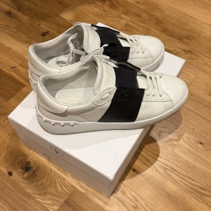 Valentino Open sneakers med svart detalj - Säljer ett par vita Valentino Open sneakers med bred svart läderdetalj över mitten. Skorna har snörning, rund tå och platt sula. Klassisk och stilren design med diskret Valentino-logga på plösen.