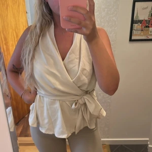 Vit omlottopp med knytband - Säljer en snygg vit omlottopp med knytband i midjan och volang nertill. Toppen har en djup v-ringning och är ärmlös, perfekt för en fräsch och stilren look.