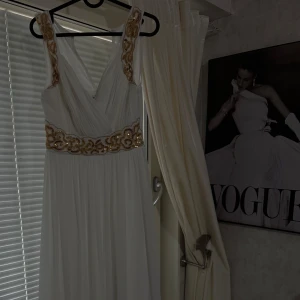 Vit långklänning med guldpaljetter - Elegant vit långklänning med v-ringad omlottringning och breda axelband. Klänningen har vackra guldpaljetter och broderier längs midjan och axelbanden. Plisserad kjol ger ett luftigt och lyxigt intryck. Perfekt för bal eller fest.