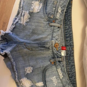 Ljusblå jeansshorts från H&M Divided - Säljer ett par ljusblå jeansshorts från H&M Divided med slitna detaljer och fransig kant. De är Lågmidjade och har 39cm i midjemått 