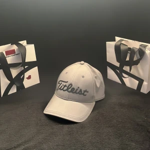 Ljusgrå keps från Titleist - Snygg ljusgrå keps från Titleist med broderad logga framtill. Klassisk sexpanelsdesign med böjd skärm och justerbar passform. Perfekt för golfbanan eller som stilren accessoar.