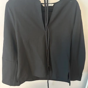 Svart kavaj med knyt från Zara - Stilren svart kavaj från Zara med knytband framtill och dekorativa knappar vid ärmslut. Kavajen har lång ärm och slits i sidorna, perfekt för en trendig och enkel look. Man knyter bandet i midjan. Saknar en boll på ena sidan av armen