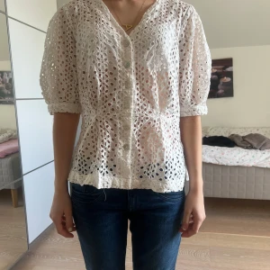 Vit blus med broderade hål och puffärmar - Säljer en söt vit blus med broderade hål och knappar framtill. Blusen har korta puffärmar och v-ringning. Perfekt för vår och sommar, lätt och luftig i bomull.
