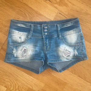 Lågmidjade jeansshorts - Snygga blå jeansshorts med slitningar framtill, pärldetaljer och dekorativa knappar. Ena bakfickan har vit spets med blommönster. Perfekta för sommaren och har en normal passform. Midjemått: 36cm men stretchiga! Kontakta vid frågor😇