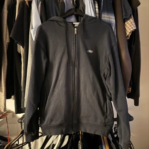 Mörkblå hoodie med dragkedja från Reebok - Säljer en mörkblå hoodie med dragkedja framtill och huva. Tröjan har en liten Umbro-logga på bröstet och ribbade muddar vid ärmslut och nederkant. Perfekt för en avslappnad stil.