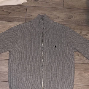 Grå ribbad kofta från Polo Ralph Lauren - Säljer en grå ribbstickad kofta från Polo Ralph Lauren med dragkedja framtill och hög krage. Klassisk logga broderad på bröstet. Perfekt för lager-på-lager och enkel att matcha med det mesta.