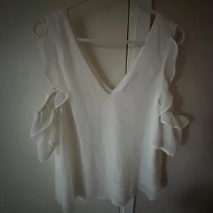 Vit blus med volangdetaljer och v-ringning - Säljer en vit blus med djup v-ringning och volangdetaljer längs ärmarna. Blusen har en luftig och flowy passform och är perfekt för en stilren look. Ärmarna är korta med öppna partier och volanger.