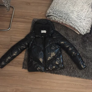 Svart pufferjacka från Calvin Klein Jeans - Säljer en svart glansig pufferjacka från Calvin Klein Jeans. Jackan har huva, dragkedja framtill och en modern, vadderad look. Perfekt för kyligare dagar och riktigt snygg till streetwear-stil. Köpt för förra vintern på kidsbrndstore för 2000 kronor.