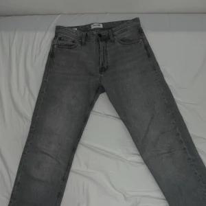 Grå jeans från Jack & Jones - Säljer ett par grå jeans från Jack & Jones i modellen CHRIS. Klassisk femficksdesign med dragkedja och knapp. Jeansen har en rak passform och är perfekta för en avslappnad stil.