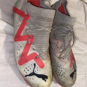 Gråa fotbollsskor från Puma med röd detalj - Säljer ett par gråa fotbollsskor från Puma med röda och svarta detaljer. Skorna har stickad strumpliknande öppning, snörning och perforerade partier för ventilation. Puma-logga på tån och sidan. Perfekta för dig som vill sticka ut på planen.