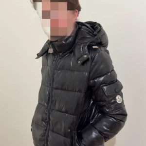Svart dunjacka från Moncler - Säljer en svart, glansig dunjacka från Moncler med huva och dragkedja. Jackan har lång ärm, quiltad design och logotyp på ärmen. Perfekt för kalla dagar och har en klassisk pufferstil.