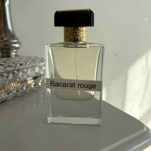 Baccarat Rouge parfym - Elegant parfymflaska med rak, transparent design och svart fyrkantigt lock med gulddetaljer. Innehåller en ljusgul vätska och etiketten 'Bacarat rouge' på framsidan. Perfekt för dig som gillar lyxiga dofter.