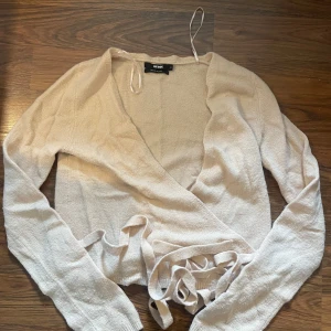Beige omlottkofta - Säljer en beige omlottopp från NA-KD med långa ärmar och knytband i midjan. Tunn och mjuk kvalitet. Snygg v-ringning och avslappnad passform.