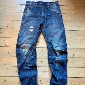 Blå slitna jeans från G-Star RAW - Säljer ett par blå jeans från G-Star RAW med slitningar och hål på både fram- och baksida. Klassisk femficksmodell med dragkedja och knapp. Jeansen har en rak passform och tydliga slitdetaljer för en tuff look.