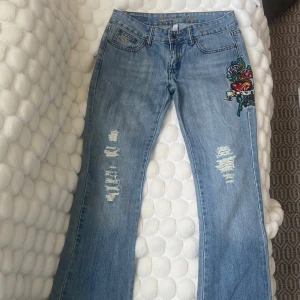 Blå bootcut jeans med broderad detalj - Snygga blå bootcut jeans med slitningar och broderad tatuering-inspirerad detalj på ena benet. Klassisk femficksmodell och låg midja. Perfekta för dig som vill ha en unik touch på dina jeans. Midjemåttet är ca 36 cm och innerbenslängden är cirka 82cm 💞
