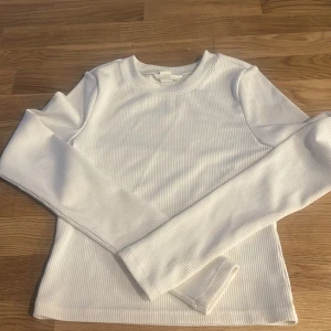 Vit ribbad långärmad tröja från H&M - Säljer en vit ribbad långärmad tröja från H&M. Tröjan har en klassisk rund halsringning och är tillverkad i ett mjukt och stretchigt material som sitter snyggt på kroppen. Perfekt basplagg till garderoben!