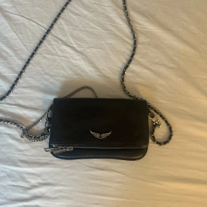 Svart axelväska från Zadig & Voltaire - Säljer en svart axelväska från Zadig & Voltaire i skinn med silverfärgad kedjerem och dragkedjedetalj framtill. Väskan har en liten silvervinge som dekoration på framsidan och är perfekt för att bära det viktigaste.