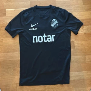 Svart AIK fotbollströja från Nike - Säljer en svart AIK fotbollströja från Nike med klubbmärke och sponsortryck på bröstet. Tröjan har korta ärmar och är tillverkad i ett lätt och ventilerande material. Perfekt för match eller träning.