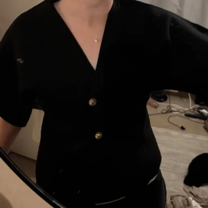 Svart kortärmad skjorta med guldknappar - Snygg svart skjorta med korta ärmar och v-ringning. Skjortan har tre dekorativa guldknappar framtill och en stilren, enkel design som passar till mycket. Perfekt för dig som gillar en clean och klassisk look.