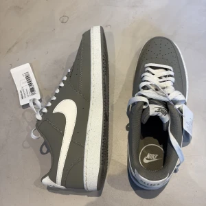 Nike Court Vision Low grå/vita sneakers - Snygga Nike Court Vision Low sneakers i grått med vita detaljer. Skorna har klassisk Nike-logga på sidan och vita skosnören. Perfekta för en stilren vardagslook.