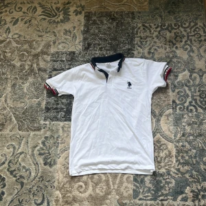 Vit pikétröja från USPA med broderad logga - Vit pikétröja från USPA med mörkblå krage och ärmslut med röd, vit och blå rand. Broderad USPA-logga på bröstet. Klassisk piké med korta ärmar och knappslå vid halsen. Storlek är jag inte säker på men denna är liten skulle säga att den passar en 8 åring pojke 