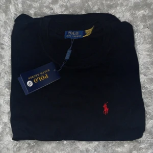 Svart t-shirt från Polo Ralph Lauren - Svart t-shirt från Polo Ralph Lauren med klassisk röd broderad logga på bröstet. T-shirten har rund halsringning och korta ärmar. Perfekt för en stilren och enkel look.