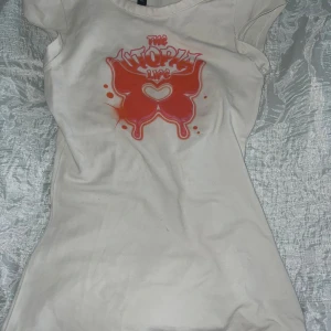 Y2K slim fit top - En vit  topp med korta ärmar och orange tryck framtill med text. T-shirten har en figurnära passform och är perfekt för dig som gillar streetstyle och y2k style