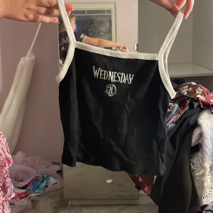 Svart ribbad linnetopp från H&M med Wednesday-tryck - Svart ribbad linnetopp från H&M med vita axelband och kant. Framtill står det 'Wednesday' i vitt tryck. Perfekt för dig som gillar serien eller vill ha en cool topp till sommaren.