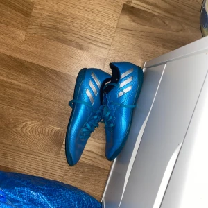 Adidas Messi 16.4 inomhus  - Säljer ett par blå fotbollsskor från Adidas med silverfärgade ränder på sidorna. Skorna har snörning och är tillverkade i syntet.perfekta för ditt barn som vill prova på fotboll eller som behöver skor till idrotten på skolan.(ena skon har ett pytte litet hål i sulan)